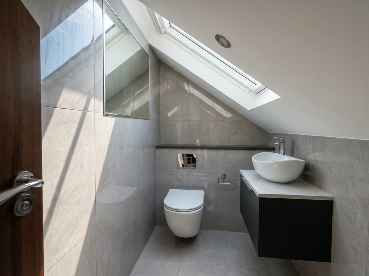 A loft en-suite shower room with a rooflight above, grey marble-effect tiling, a wall-hung WC and a vessel basin on a dark vanity unit
